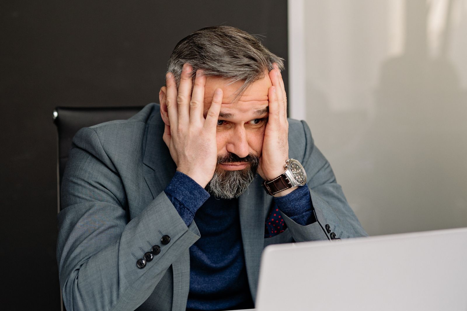 Homme à son bureau, les deux mains plaquées sur le visage, le regard figé sur l'écran de son ordinateur portable, l'air de regretter une décision