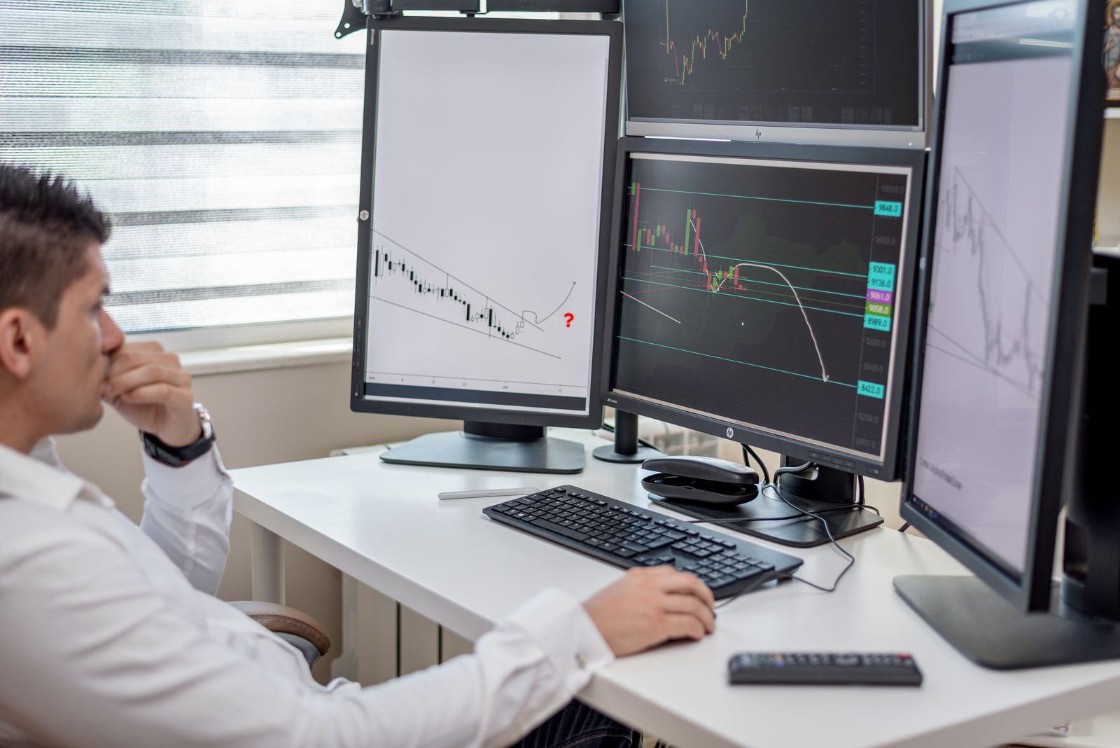Trader in a white shirt, one hand on the mouse and the other at his mouth, hesitating in front of several chart screens