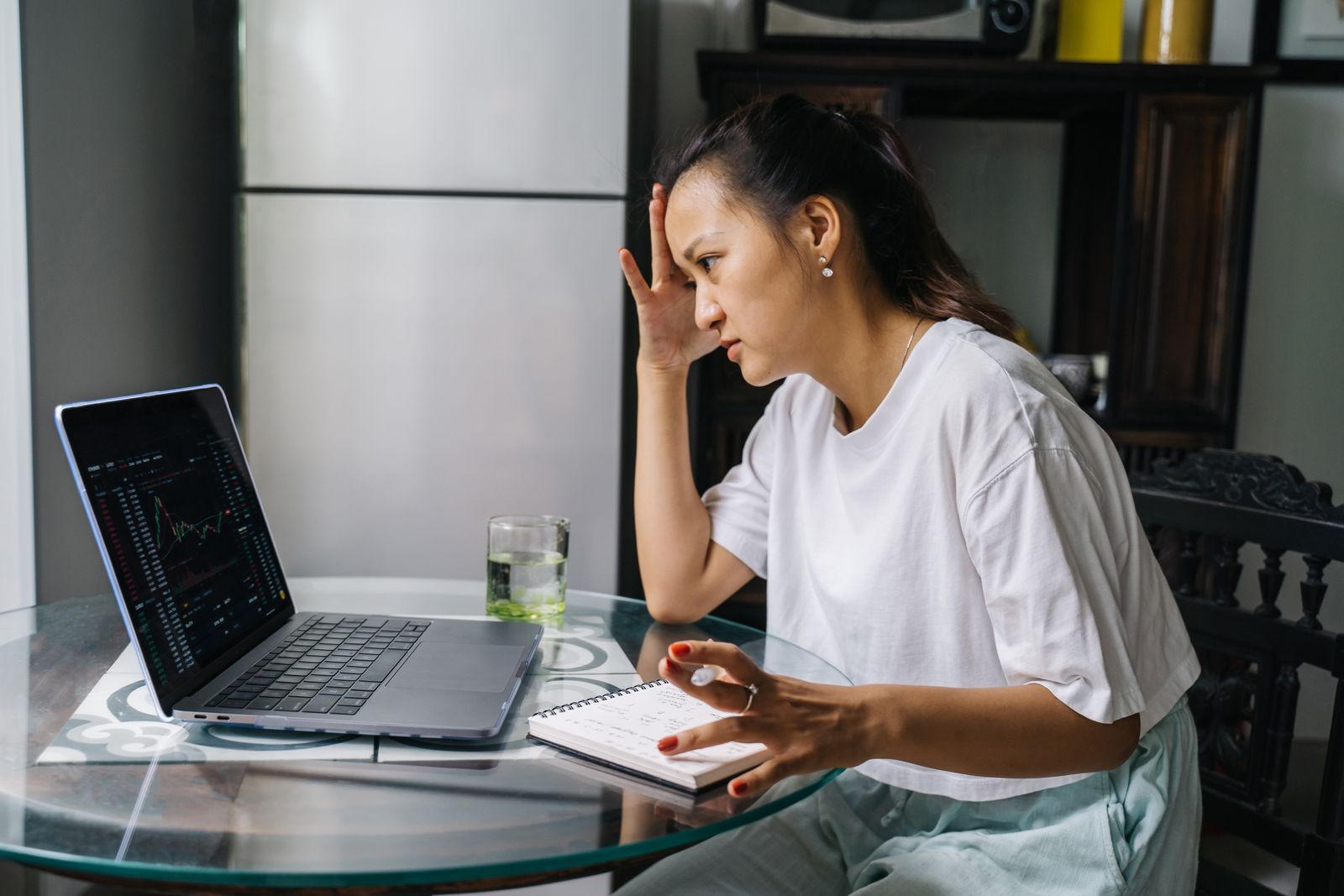 Femme qui trade depuis chez elle, une main sur le front, le regard tendu vers un graphique sur son ordinateur portable, un carnet de notes ouvert sur la table